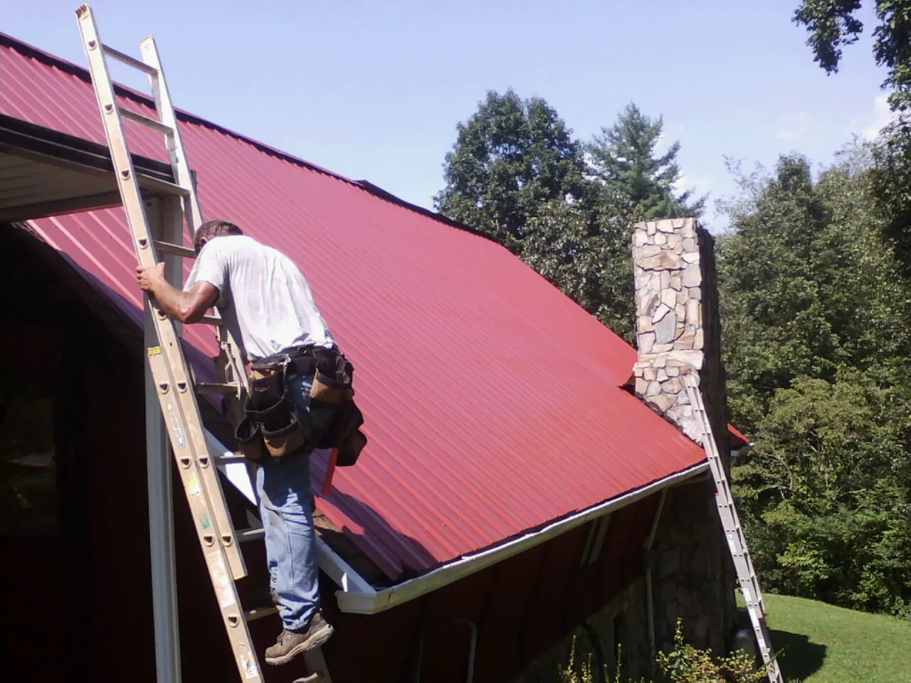 Roofer on a ladder beside a red metal roof and stone chimney — Soffit & Fascia Repair by Roofing Brothers in Saint George